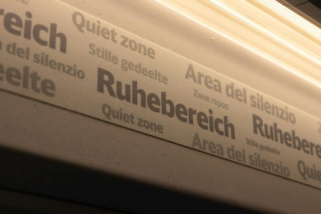 A close-up, angled view of a "Ruhebereich" (Quiet zone) sign inside a European train carriage. The textured grey panel displays text in multiple languages, including "Area del silenzio," "Zone repos," and "Stille gedeelte," illuminated by warm overhead strip lighting.