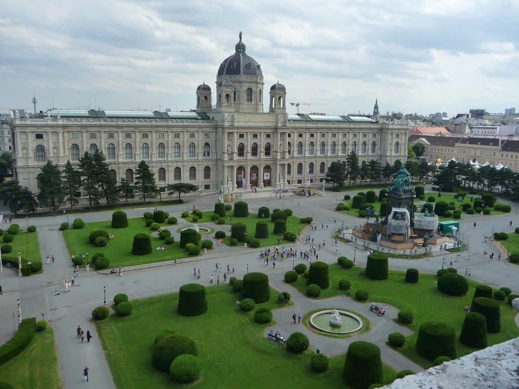 High-angle view of the Naturhistorisches Museum in Vienna, Austria, displaying grand Neo-Renaissance architecture and a central dome, overlooking the manicured topiary gardens and statues of Maria-Theresien-Platz under an overcast sky.