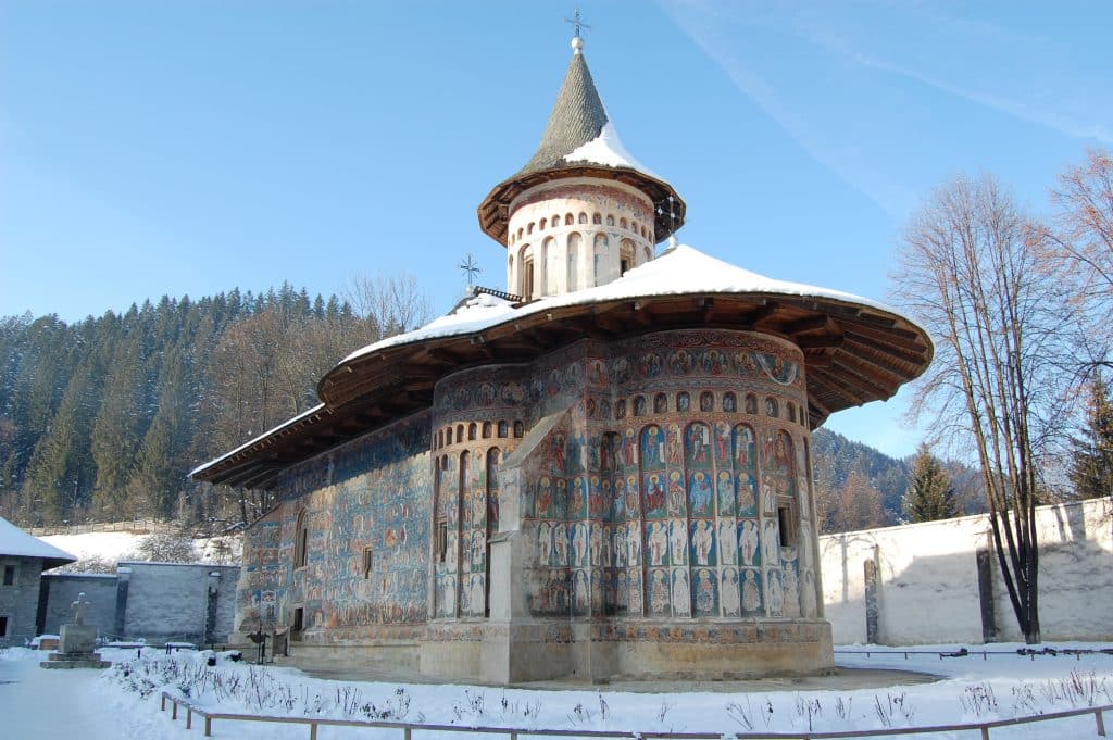 A side view of the Voroneț Monastery in Romania during winter, showcasing its famous exterior frescoes featuring "Voroneț Blue" pigments. The church has a snow-covered, wide-eaved roof and a central tower topped with a cross, set against a clear blue sky and a snowy courtyard.