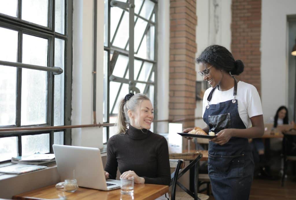 A smiling waitress in a denim apron serves a croissant on a black tray to a happy blonde customer working on a laptop at a wooden table in an industrial-style cafe. Large windows and brick walls are in the background.