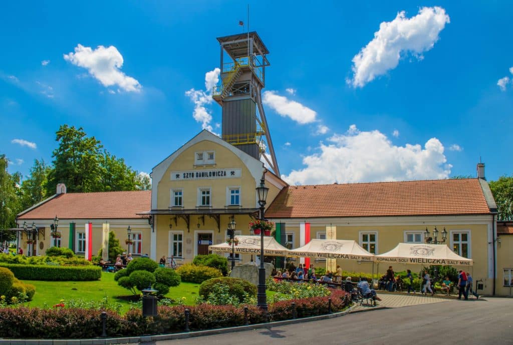 Exterior view of the Daniłowicz Shaft building at the Wieliczka Salt Mine in Poland, featuring a yellow historic building topped with a steel mining headframe. The facade displays a sign reading "Szyb Daniłowicza," and white tents labeled "Kopalnia Soli Wieliczka Salt Mine" sit near a manicured garden with tourists.