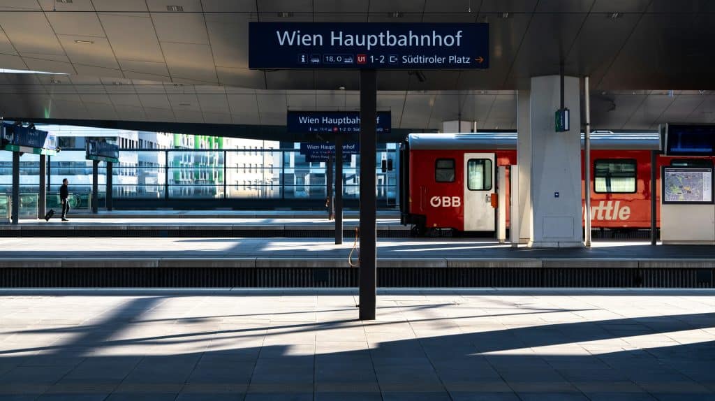 Getting Around Central & Eastern Europe: Trains, Buses, and Rental Cars - A sunny platform view at Wien Hauptbahnhof (Vienna Main Station). A dark blue sign with white text reads "Wien Hauptbahnhof" and indicates connections to U1 and Südtiroler Platz. To the right, an orange and white ÖBB Cityshuttle train is parked. On the left platform, a lone traveler pulls a rolling suitcase. Long, sharp shadows stretch across the tiled ground.