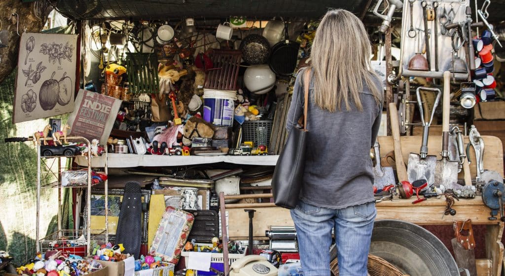 A woman seen from behind browsing a cluttered antique market stall filled with vintage tools, toys, and kitchenware. A paper bag with the text "INDIE FESTIVAL" sits on a shelf among the bric-a-brac.
