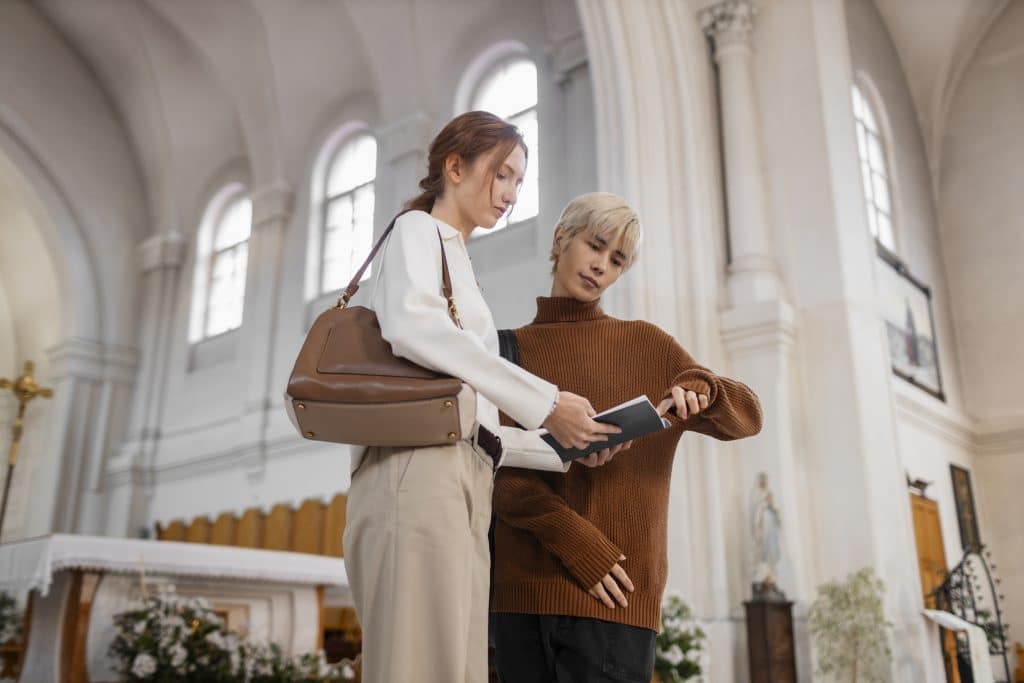 A low-angle medium shot of two young visitors standing inside a bright church with high white arches. A woman with reddish-brown hair holds a dark brochure while a blonde man in a brown turtleneck sweater points at the text. In the background, a blurred altar, a gold crucifix, and large arched windows are visible.