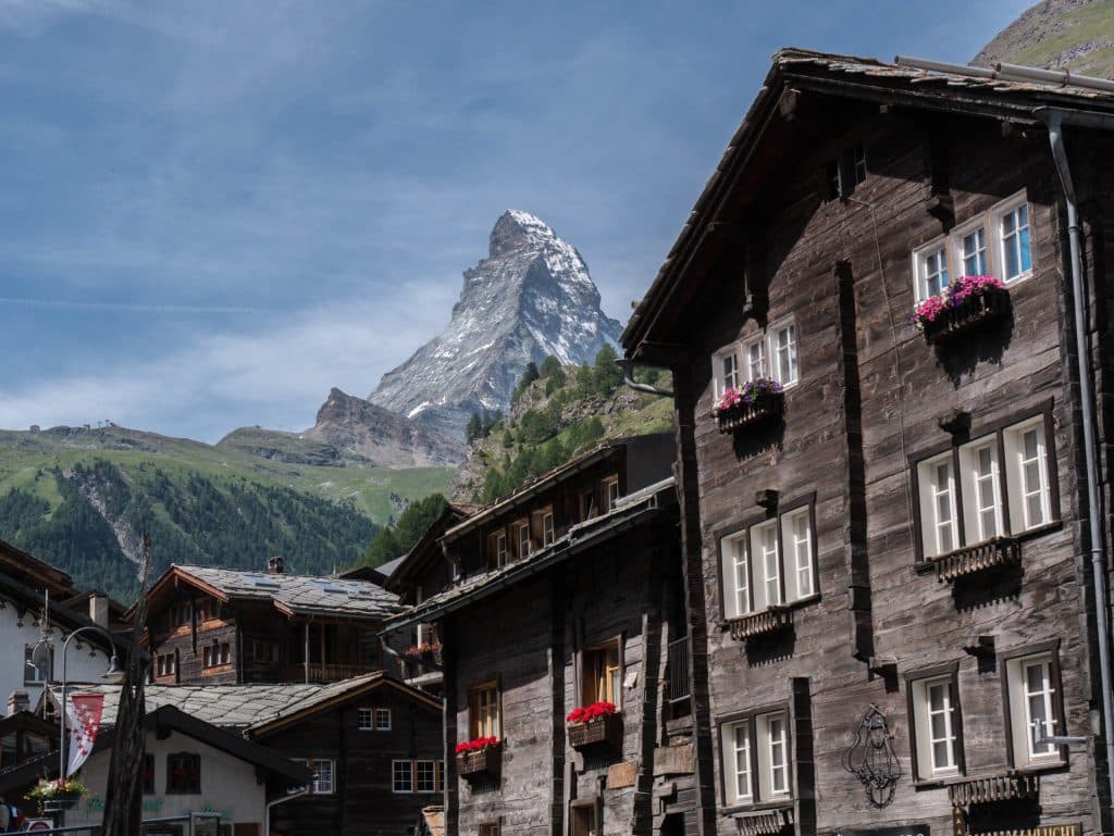 A view of traditional dark wood Swiss chalets with flower boxes in Zermatt, framing the Matterhorn rising in the background under a blue sky.