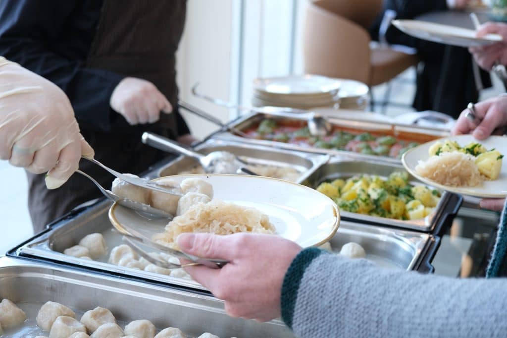 A close-up of a person serving boiled pierogi and sauerkraut onto a plate from a cafeteria-style buffet.