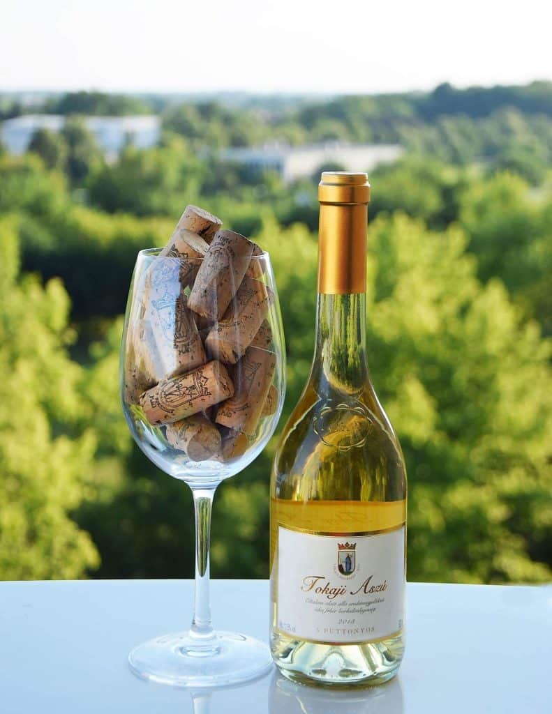 A bottle of golden Tokaji Aszú wine standing next to a wine glass filled with corks, with a vineyard view in the background.