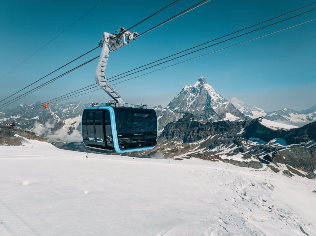 modern, sleek blue and black cable car gondola suspended high on cables over a white snowy glacier field, with the Matterhorn peak in the distance.
