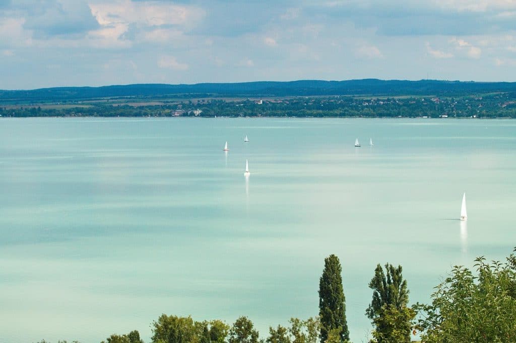 Wide panoramic view of Lake Balaton's turquoise water with sailboats in the distance and hills on the horizon.