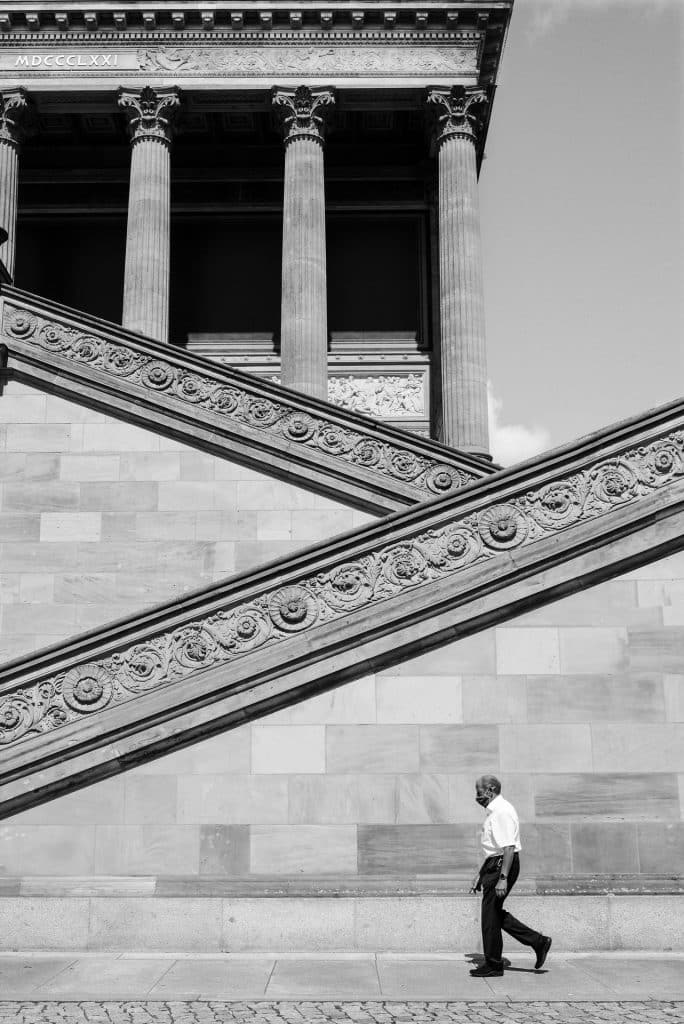 A black and white photograph of the grand staircase and Corinthian columns of the Alte Nationalgalerie in Berlin. Roman numerals MDCCCLXXI are inscribed on the building. A lone man walks in profile at the bottom right.