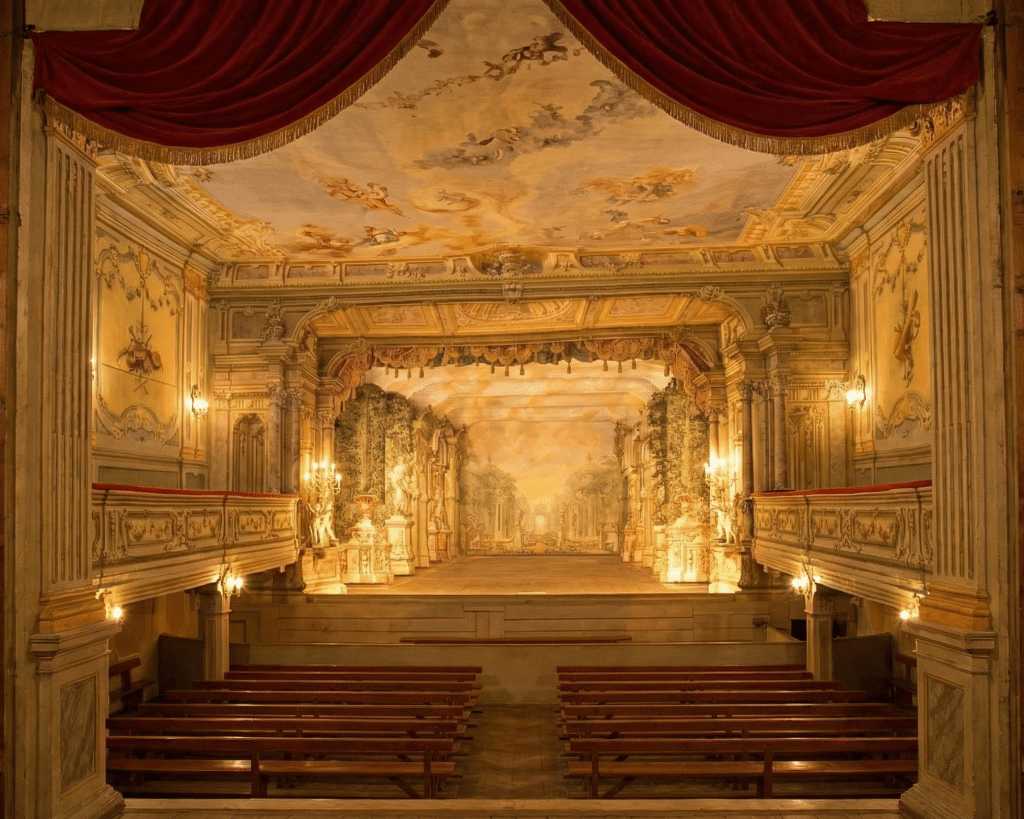 Interior view of the Baroque Castle Theater in Český Krumlov showing the painted stage set with forced perspective and empty wooden benches.