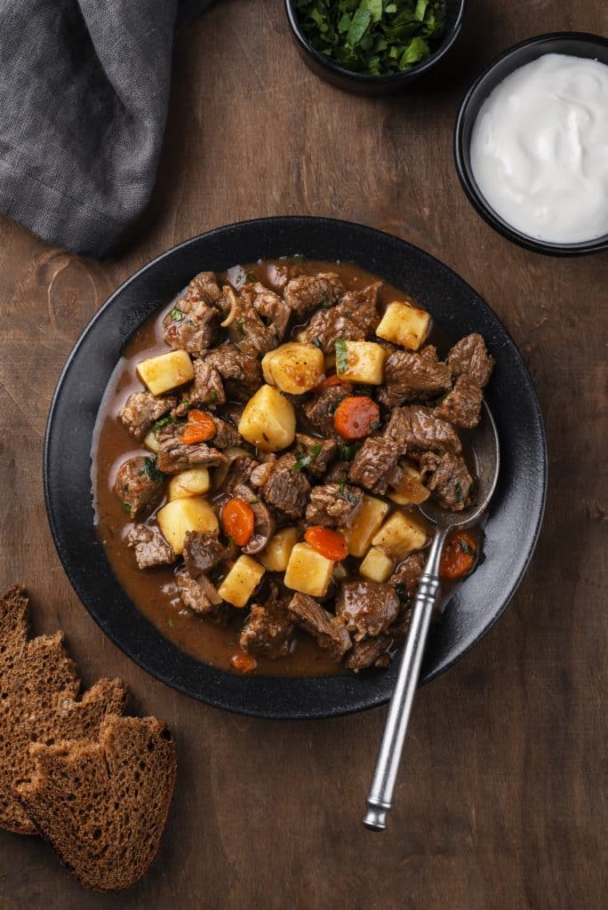 Top-down view of a thick, hearty beef stew with potatoes and carrots served in a black bowl with sour cream on the side.