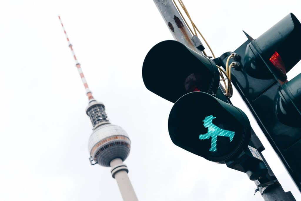 berlin first time visitor - A low-angle shot of a green Ampelmann traffic light signal in focus. The Berlin TV Tower (Fernsehturm) is visible in the background, blurred by shallow depth of field, against a white overcast sky.