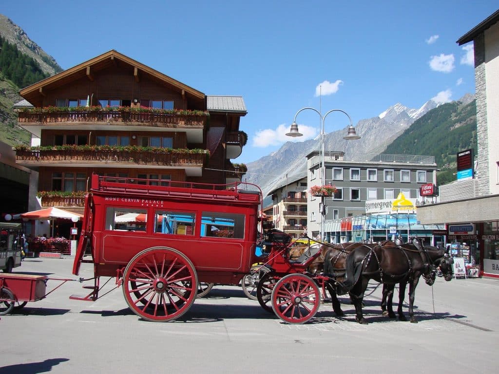 A red horse-drawn carriage from the Mont Cervin Palace waiting on a sunny street in Zermatt with traditional buildings and mountains in the background.