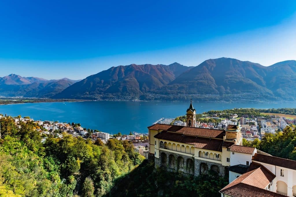 High-angle panoramic view of the Madonna del Sasso sanctuary perched on a cliff above Locarno, overlooking Lake Maggiore and the Swiss Alps.