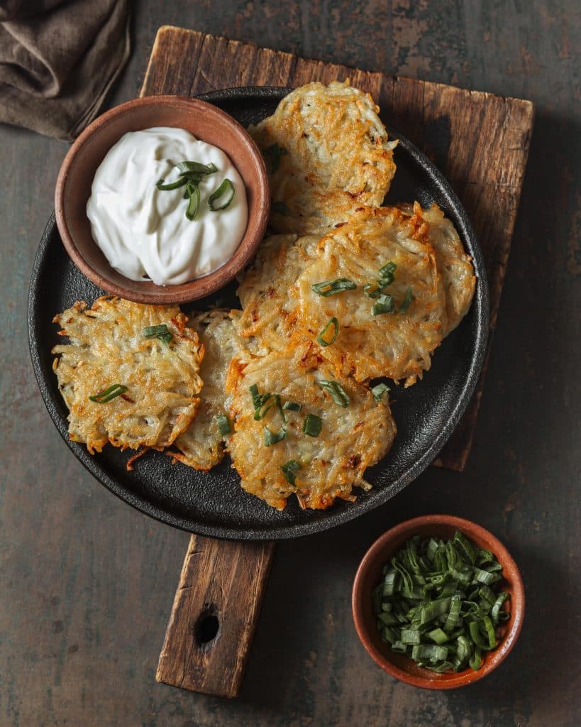 Overhead shot of golden-brown potato fritters (Rösti or Latkes) served in a black cast iron skillet with a side of sour cream and chopped green onions.