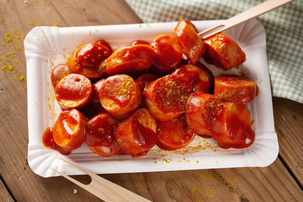 A top-down close-up of Currywurst served on a white paper plate. The sliced sausage is covered in red ketchup and sprinkled with yellow curry powder. A wooden fork rests on the side, set against a wooden table background.