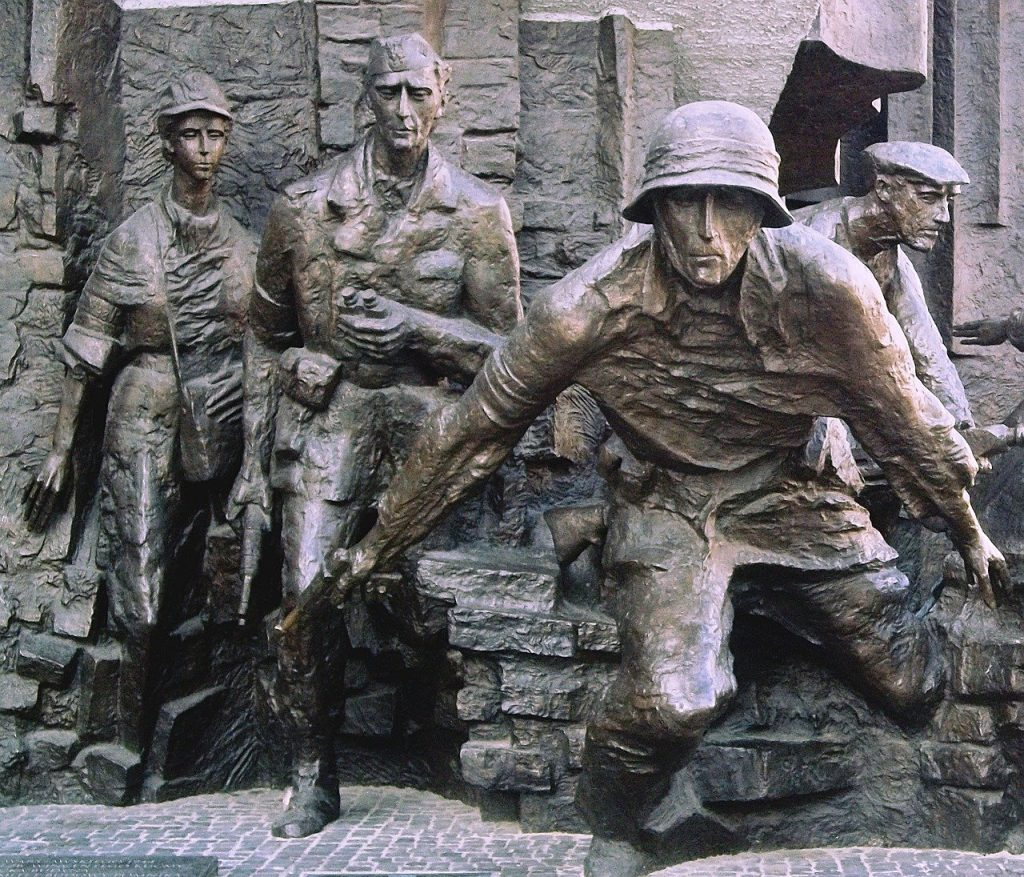 A close-up of bronze statues from the Warsaw Uprising Monument, depicting soldiers and civilians emerging from stone blocks, faces tense with determination.