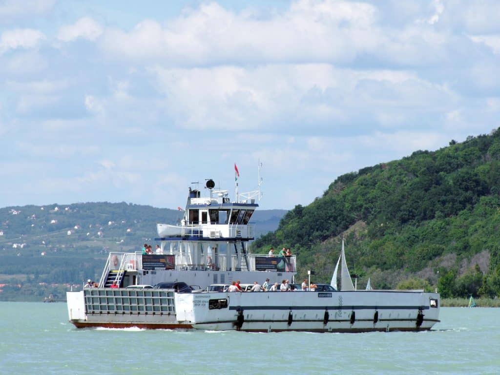 A white ferry boat loaded with cars and passengers crossing the turquoise waters of Lake Balaton towards the green hills of Tihany.