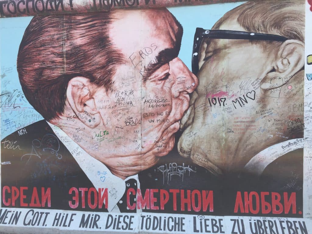 A close-up photograph of the famous "Fraternal Kiss" mural on the Berlin Wall's East Side Gallery, depicting Leonid Brezhnev and Erich Honecker kissing. The artwork is covered in visitor graffiti signatures and tags. The text at the bottom reads "MEIN GOTT, HILF MIR, DIESE TÖDLICHE LIEBE ZU ÜBERLEBEN".