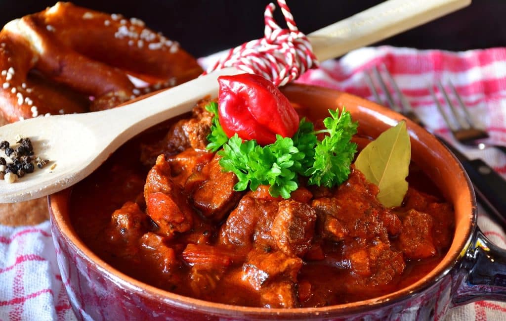 A rustic red ceramic pot filled with rich beef goulash soup garnished with fresh parsley, sitting next to a wooden spoon and pretzels.