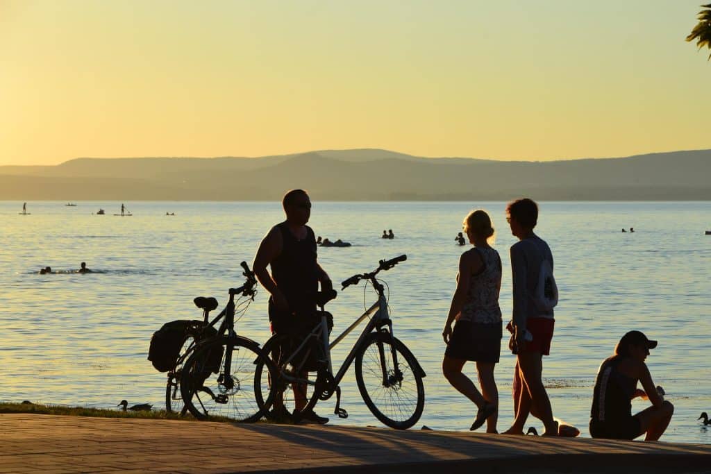 Silhouettes of cyclists and people standing by Lake Balaton at sunset with golden light reflecting on the water.