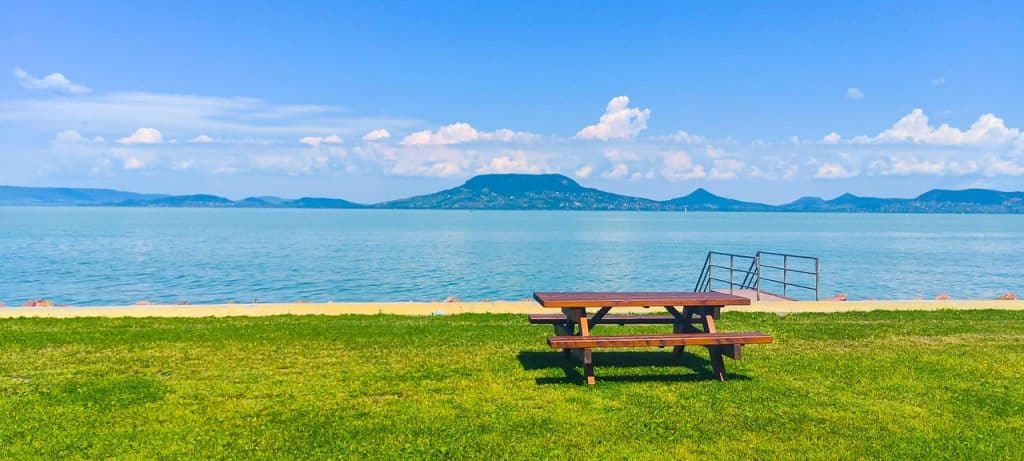 A wooden picnic table on a grassy beach on the South Shore looking out over turquoise water toward the flat-topped Badacsony volcanic hill.