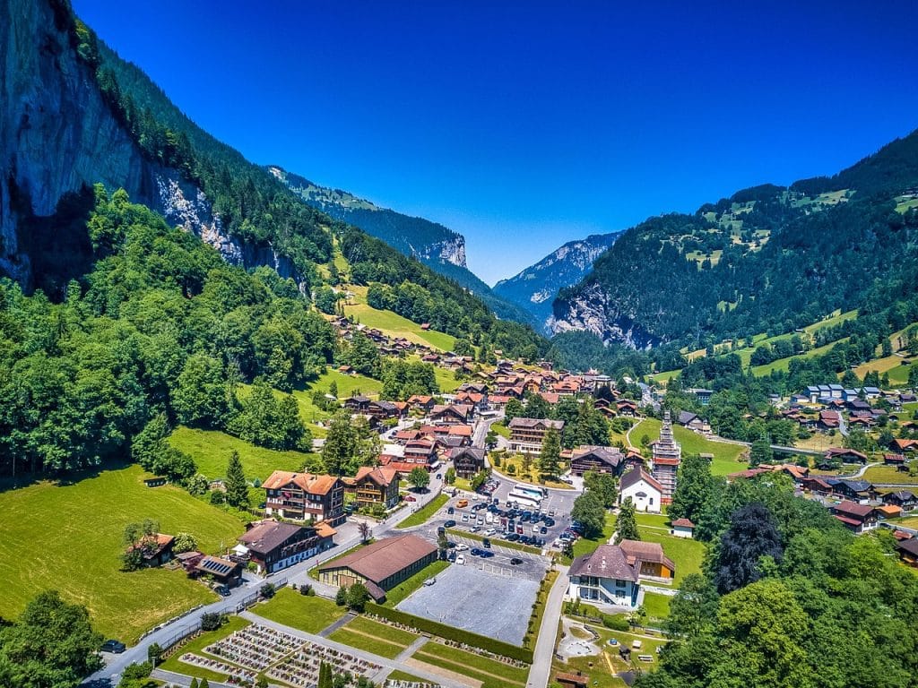 High-angle aerial view of the Lauterbrunnen valley village, featuring the Staubbach Fall, traditional chalets, and lush green cliffs against a blue sky.