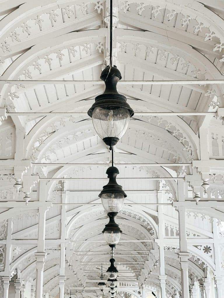 A low-angle view looking up at the intricate white wooden ceiling and arches of the Market Colonnade in Karlovy Vary, featuring a row of vintage black hanging streetlamps