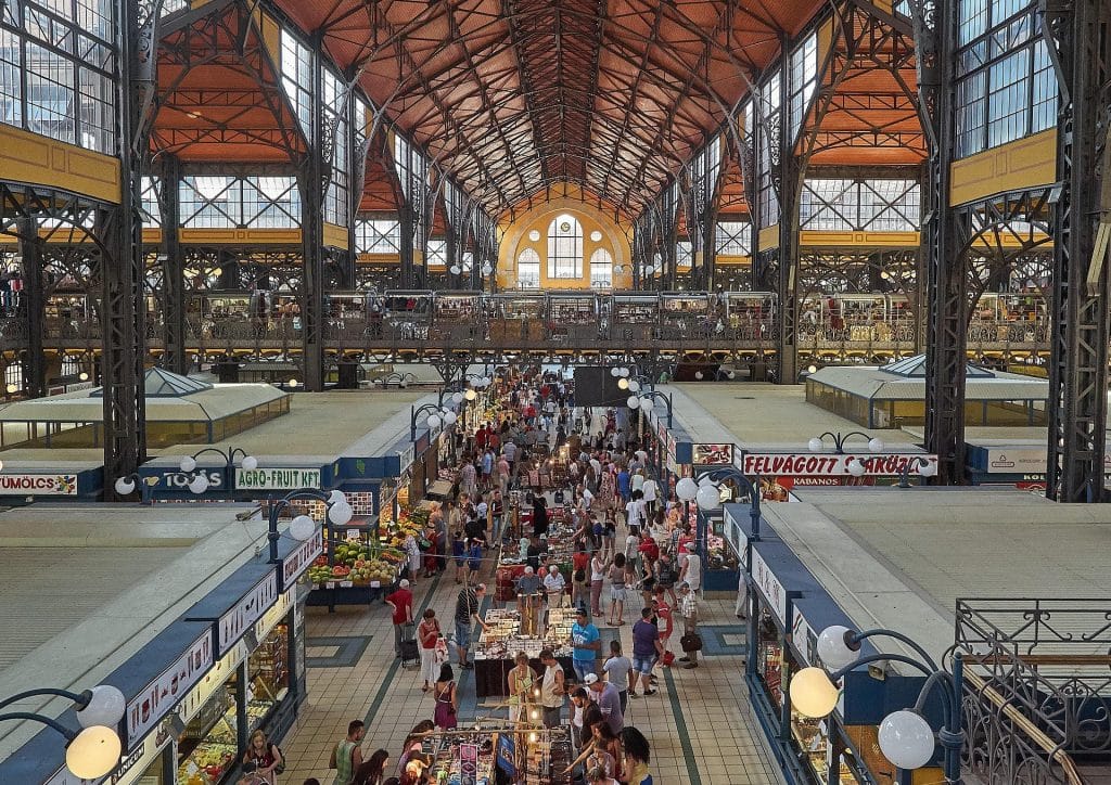 A wide interior shot of the Budapest Central Market Hall, showing the high steel roof and crowds of people shopping at stalls.