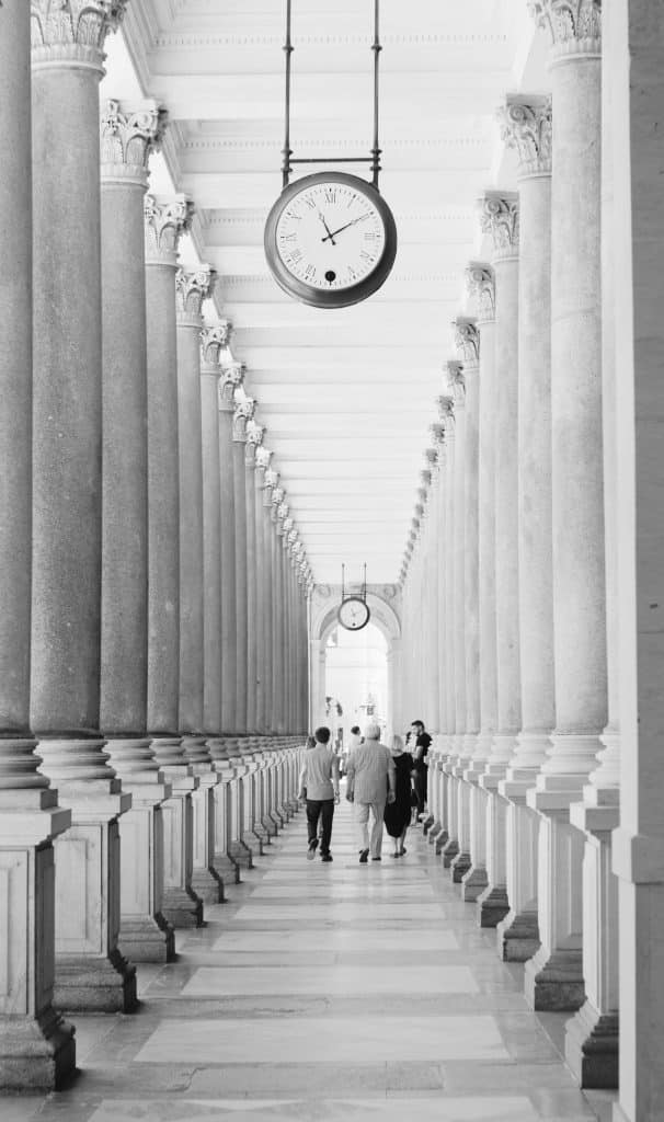 Black and white photography of people walking slowly through the Mill Colonnade in Karlovy Vary. Caption: The "walking cure" in the Mill Colonnade: A ritual unchanged since the 19th century.