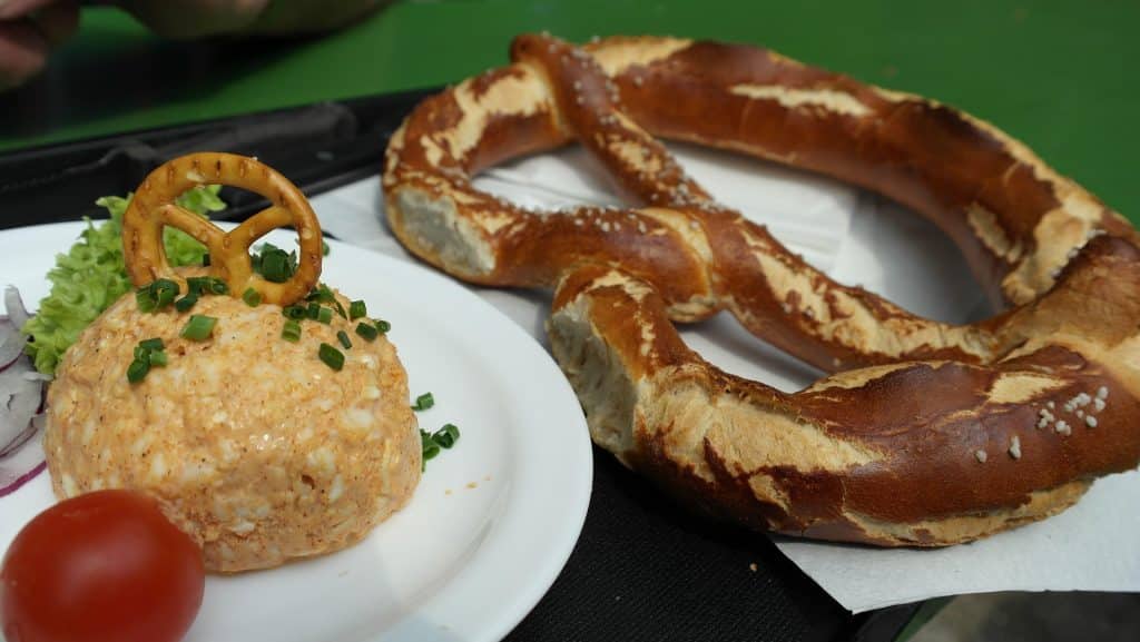 A scoop of Obatzda cheese spread garnished with chives and a mini pretzel, served on a white plate next to a large fresh Bavarian soft pretzel.