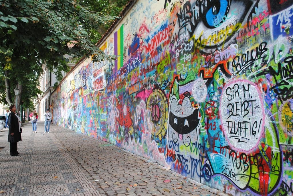 A colorful section of the John Lennon Wall in Prague covered in layers of graffiti, lyrics, and street art. A tourist stands to the left admiring the wall. Text visible includes 'Sir M. Bleibt Fluffig'.