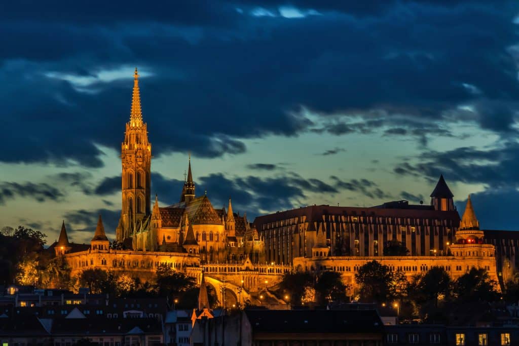Night view of the illuminated Matthias Church spire and Fisherman's Bastion fortifications against a dark blue sky.