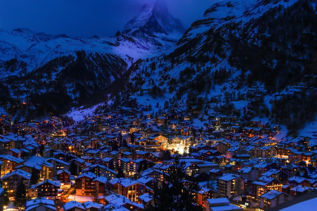 A high-angle view of Zermatt village at night (Blue Hour), with warm lights glowing from the dense chalet rooftops and the dark triangular silhouette of the Matterhorn looming behind.
