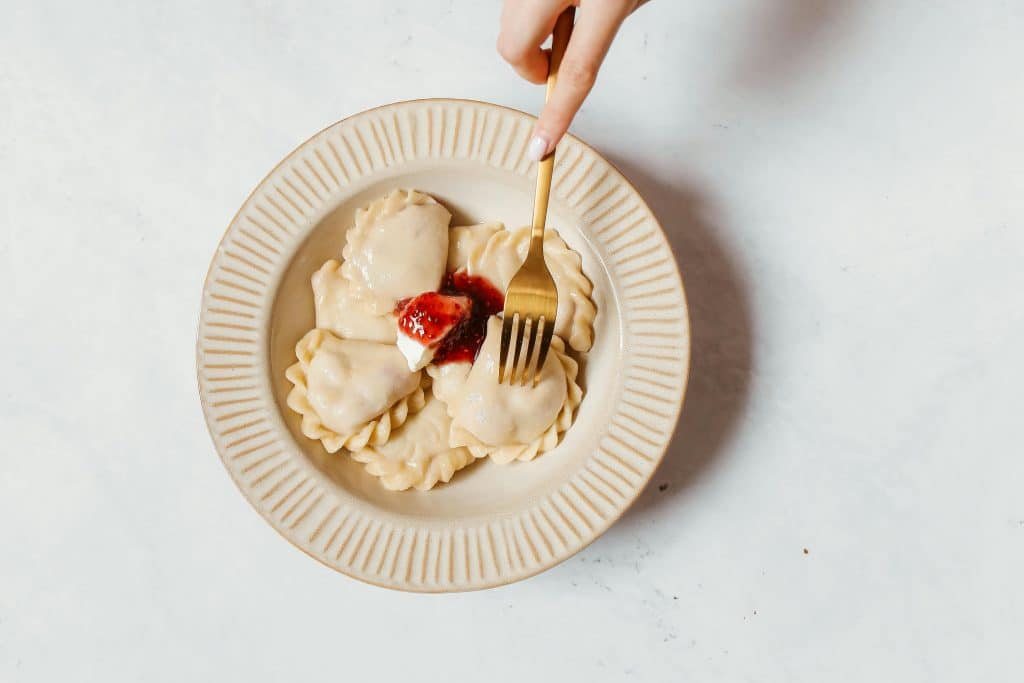 Top-down view of a ceramic plate with dumplings (pierogi) topped with sour cream and red berry jam; a hand holds a gold fork pressing into one dumpling.