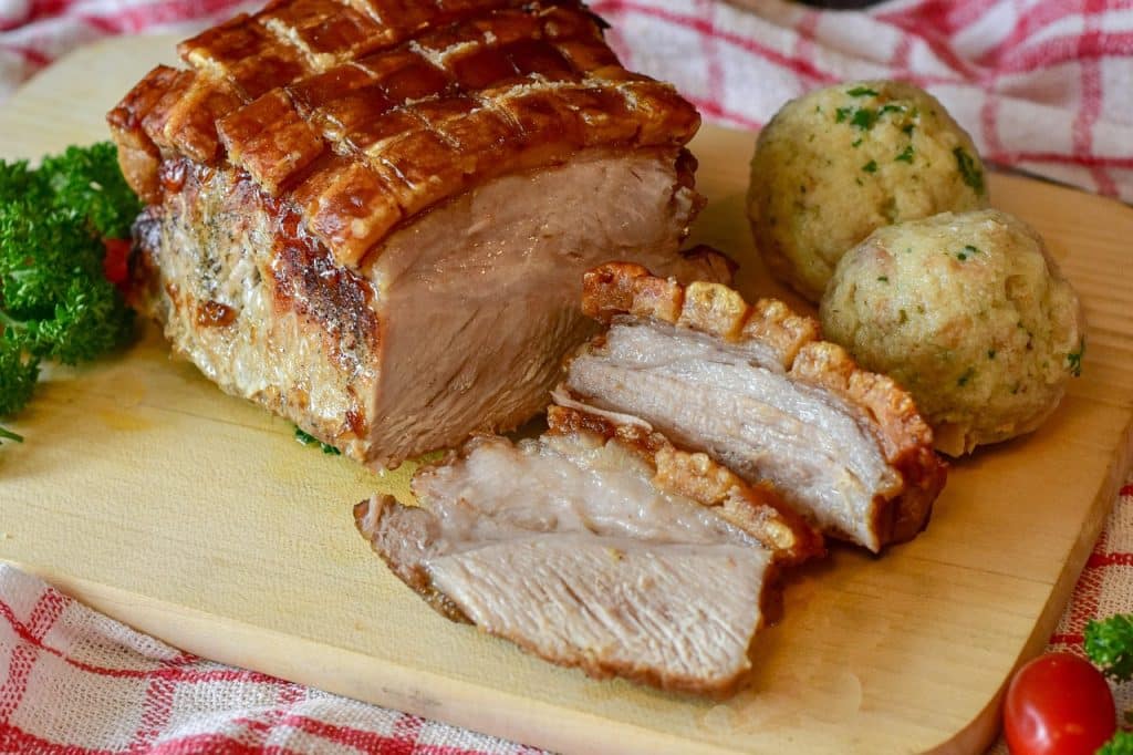 Slices of roasted pork with crispy crackling served on a wooden board with two potato dumplings and fresh parsley.