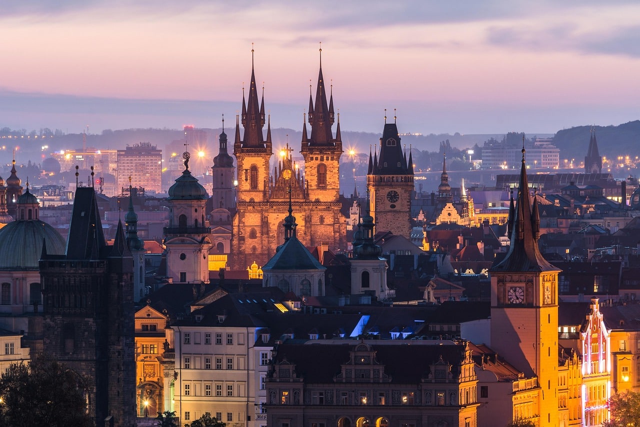Panoramic view of the Prague skyline at twilight, featuring the illuminated spires of the Church of Our Lady before Týn rising above layers of historic buildings and rooftops.