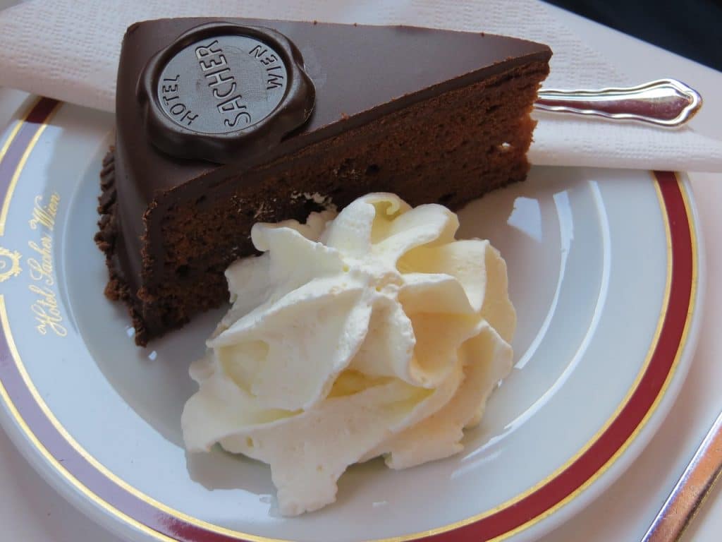 A dense chocolate sponge cake with a dark glaze, featuring the Hotel Sacher chocolate seal and a side of unsweetened whipped cream.