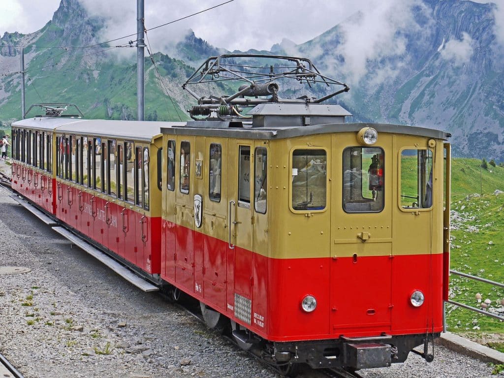 The historic red and cream Schynige Platte Bahn cogwheel train parked on a gravel track with rocky alpine mountains in the background. Side text reads "SCHYNIGE PLATTE BAHN".