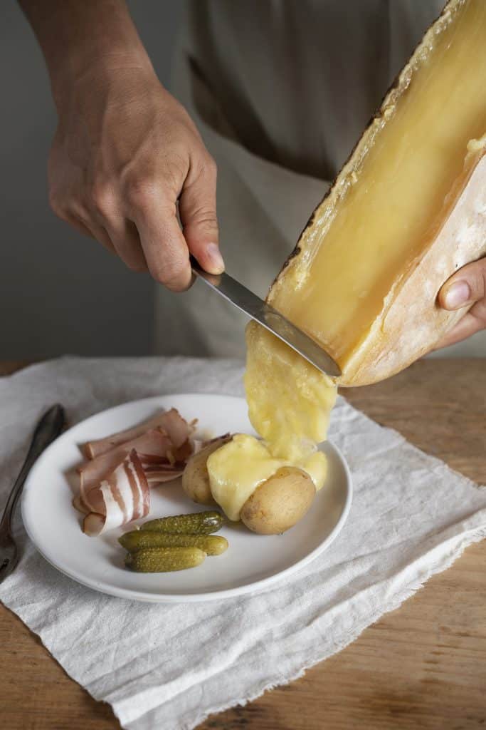 High-angle shot of a knife scraping a cascading layer of melted raclette cheese from a wheel onto a plate of boiled potatoes and cured meats.