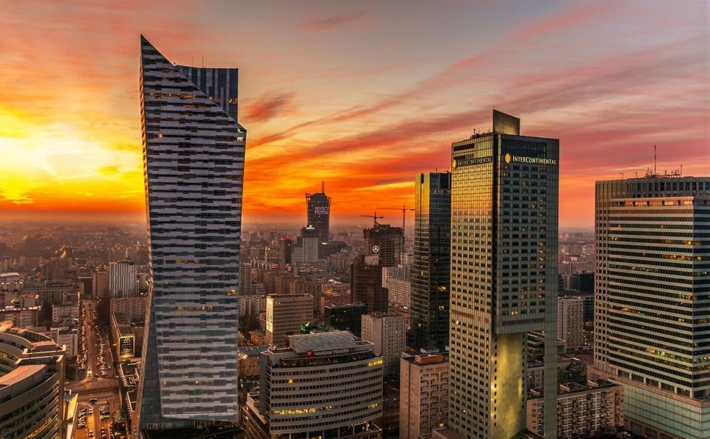A dramatic aerial view of the Warsaw skyline at sunset, featuring the curved Złota 44 tower and the InterContinental hotel glowing against a fiery orange sky.