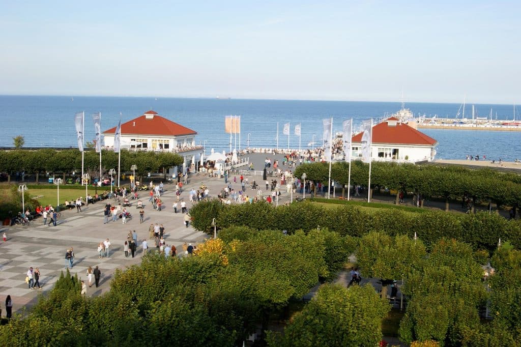 High-angle view of the Sopot coastline on a sunny day, showing a busy paved square, red-roofed spa buildings, and a long wooden pier extending far out into the calm blue Baltic Sea.