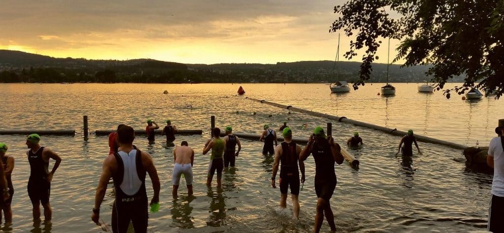 Silhouettes of swimmers entering Lake Zurich at sunrise, with sailboats and hills in the distance.