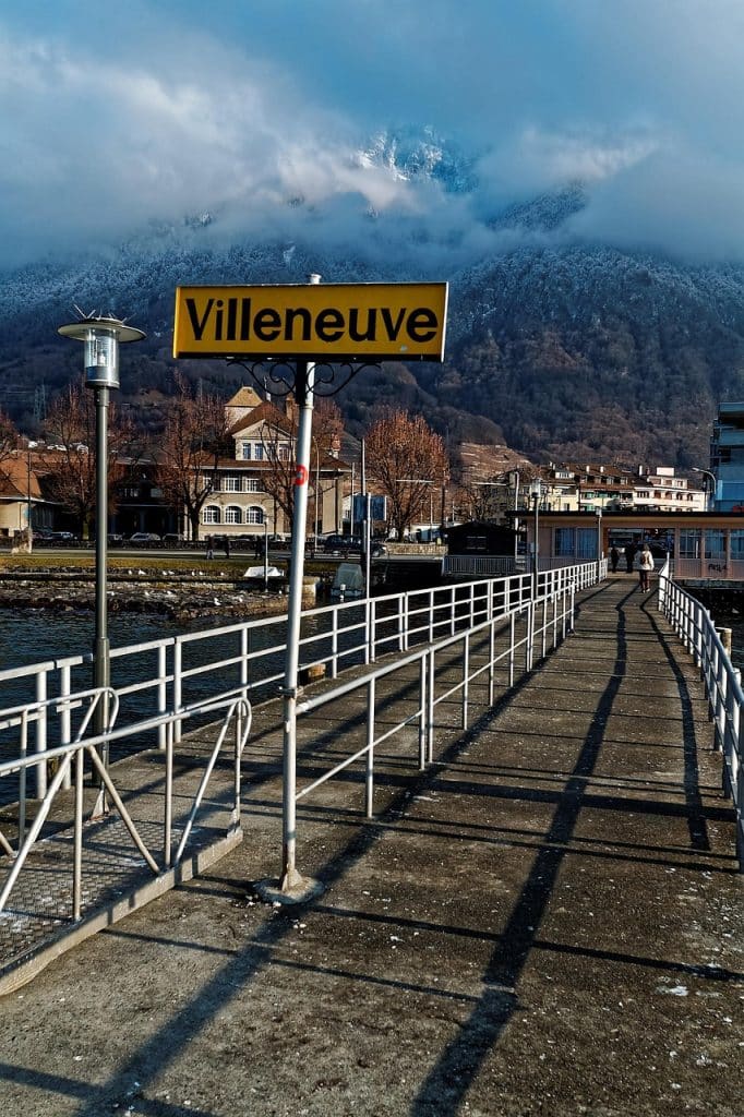 A yellow station sign reading "Villeneuve" on a pier, with moody clouds and mountains looming in the background.