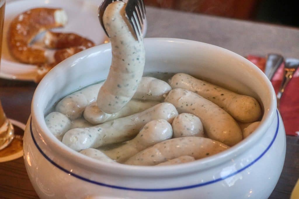 Traditional Bavarian breakfast showing white veal sausages (Weißwurst) floating in a white porcelain tureen of hot water, served with sweet mustard and pretzels.
