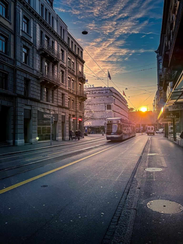 A Zurich tram moving through a city street at sunset, with golden light reflecting off the rails and buildings